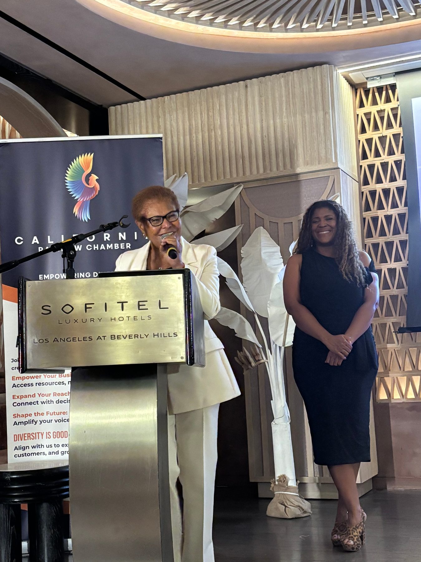 A speaker at the Sofitel podium beside a California Rainbow Chamber banner